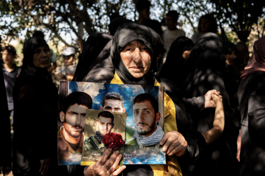 A woman holds several portraits of young men; she stands in front of a crowd of shadowed figures.