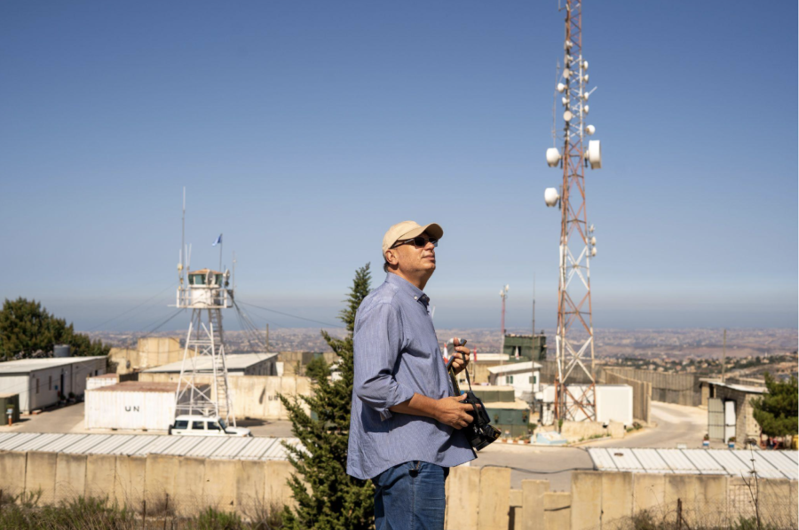 A man in a hat and a light blue shirt stands in front of a town with low buildings.
